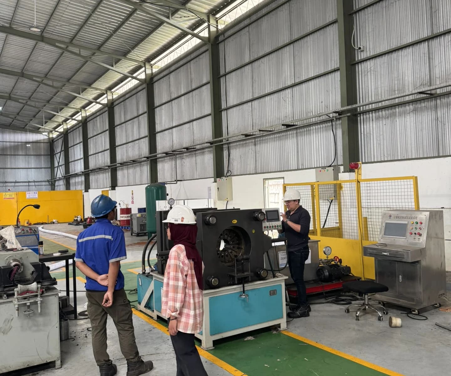 Tiga pekerja mengenakan helm keselamatan berdiskusi di depan mesin uji baja di gudang industri
